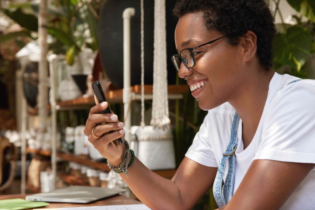 Positive dark skinned woman excited with good news getting notification on smart phone, rejoices recieving discount or credit in banking, wears white t shirt and optical glasses, sits indoor.